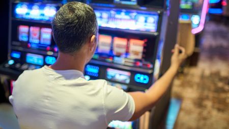 A view over the shoulder of a man wearing a white t-shirt with gray hair sitting at a slot machine