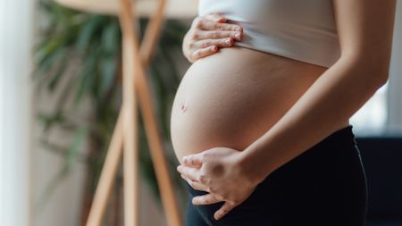 A close up of a pregnant belly, with a woman wearing a white shirt that is bunched up above it and black pants below. A lamp is blurry behind the woman