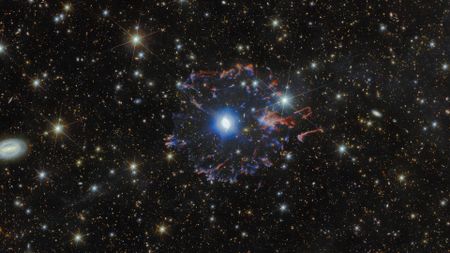 A view of the Cat's Eye Nebula, a blue ball of light surrounded by purple and red gas in a starry night sky